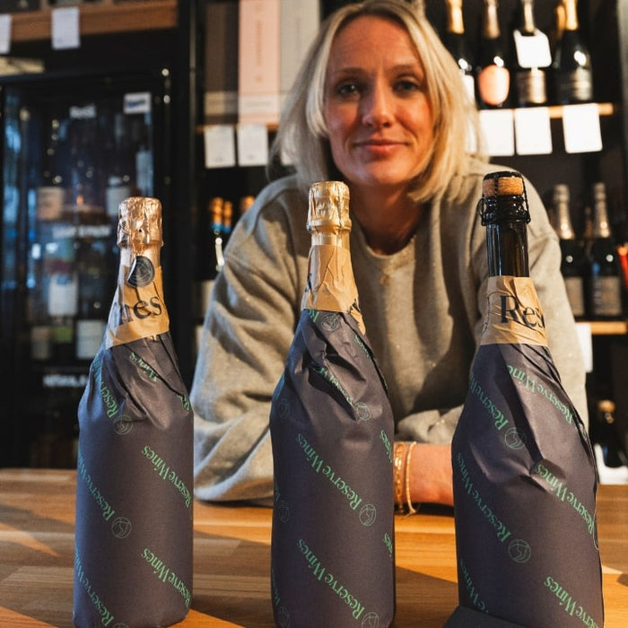 Kate Goodman behind a blind wine tasting kit - three bottles of sparkling wine in branded sleeves with a blurred bar setting.