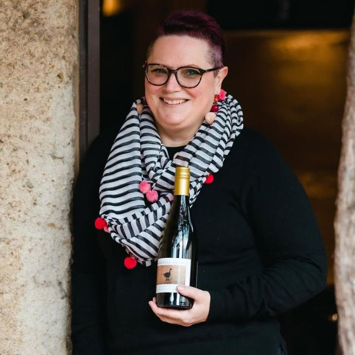 Liz Forster holding a bottle of wine with a striped scarf, standing against a stone wall.