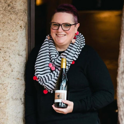 Liz Forster holding a bottle of wine with a striped scarf, standing against a stone wall.